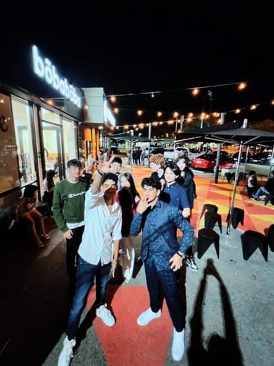 The image depicts a nighttime outdoor scene at the bōbabōba establishment located at Avenue de la Saline 24, 1420 Braine-l'Alleud, Belgium. Approximately ten young individuals, identified as teenagers or young adults, are positioned in the midground and foreground of a paved outdoor area.
The ground features a distinctive pattern of red, orange, and yellow geometric shapes against a grey surface. Long, dark shadows of the individuals are cast across this patterned ground, extending towards the lower left.
In the immediate foreground, two males are posing. The male on the left, with dark hair, wears a white long-sleeved button-up shirt, dark jeans, and white sneakers; his right hand is raised, forming a peace sign, partially obscuring his face. The male on the right, with dark hair, wears a dark blue patterned long-sleeved shirt, dark plaid pants, and white sneakers; his left hand is raised, forming a peace sign near his chin. Behind these two, a male in a dark green "Champion" sweatshirt and dark pants stands to the left. The remaining individuals in the group are positioned behind and around these three, with varied gazes, some directed towards the camera.
To the right of the foreground group, several small, dark-colored, geometric stools are arranged on the patterned ground. String lights with illuminated bulbs are suspended overhead.
The background features the brightly lit "bōbabōba" storefront, displaying its name in illuminated white letters above large glass windows revealing interior lighting and patrons. Beyond the storefront, a parking lot is visible, containing several parked vehicles, including a red sedan, and additional individuals. The scene is illuminated by artificial lighting sources against a dark night sky.