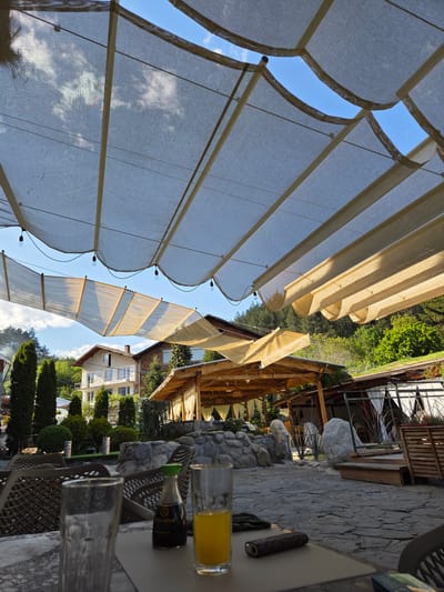 The scene is an outdoor restaurant or cafe in Slokoshtitsa, Bulgaria. The perspective is from a table looking up at canvas awnings that provide shade. Blue sky is visible through the awning. The restaurant setting includes tables and chairs, part of a stone wall, greenery, and buildings in the background. On the table are glasses, a bottle of soy sauce, and a cigar.