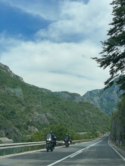 The image shows a winding road in Donji Jasenjani, Bosnia and Herzegovina. The road is bordered by a metal guardrail and a steep, green mountainside. Two motorcycles, each with two riders, are traveling on the road. The sky is cloudy with patches of blue. Part of a tree is visible in the upper left corner.