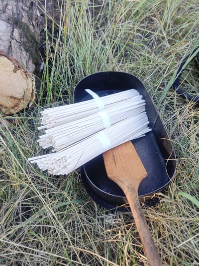 A black, rectangular pan containing several bundles of white noodles rests on dry, green grass. The noodles are tied together with white paper bands. A wooden spatula with a wide, flat head lies across the pan. To the left of the pan is a cut log, showing its rough texture and a small patch of green moss. The background consists of more dry grass and some vegetation. The lighting suggests it is daytime, possibly late afternoon, given the warm tones. The scene appears to be outdoors in a natural setting, possibly a campsite or a rural area. There are no people or animals visible. The overall mood is peaceful and perhaps preparatory, as if a meal is about to be cooked. There is no visible text in the image.