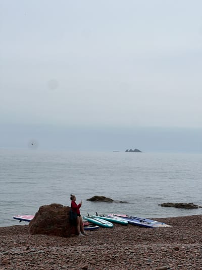 The image depicts a coastal scene in Russkiy, Russia. A woman wearing a red shirt and dark shorts sits on a rocky outcropping on a pebble-covered beach. She is facing the water and pointing with her right hand. Several paddleboards are resting on the beach next to her. The ocean stretches to the horizon under a hazy sky. A small island is visible in the distance.