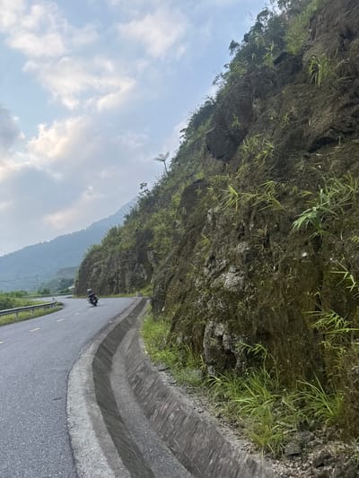 The image captures a scenic view of a paved road winding through a mountainous landscape in Tân Quang Commune, Vietnam. A dark motorcycle, with a rider, is visible on the asphalt road in the mid-ground, traveling away from the viewer. The road appears dry and has faint yellow markings on the left side, with a metal guardrail partially visible further along.

To the right of the road, a multi-tiered concrete drainage ditch or barrier separates the road from a very steep, rugged mountainside. This mountainside is primarily dark, rocky earth and stone, heavily overgrown with a variety of green vegetation, including sparse clumps of grass, ferns, and small bushes clinging to its surface. The texture of the rock and the individual plants are discernible.

In the background, beyond the immediate mountainside, multiple layers of hazy, blue-grey mountains stretch into the distance, indicating a vast and remote mountainous region. The sky occupies the upper left portion of the image, showing a mix of light blue with scattered white and grey clouds. The overall lighting suggests daytime, possibly early morning or late afternoon, with diffused light filtering through the clouds. The scene conveys a sense of peaceful travel through a natural, untamed environment. There is no visible text.