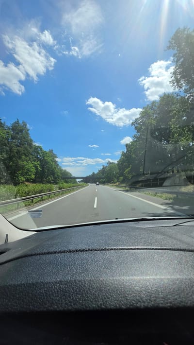 The image depicts a view from inside a vehicle, looking out through the windshield onto a two-lane road. The road appears to be a highway, bordered by a guardrail and lush green trees on both sides. The sun is shining brightly, casting rays of light that create a lens flare in the upper left corner of the image. The sky is a vibrant blue, dotted with fluffy white clouds, suggesting fair weather. The time of day is likely daytime, given the strong sunlight. The interior of the vehicle is visible on the right side of the frame, showing a textured dark gray surface, possibly part of the dashboard or door panel. A thin white line is visible on the road, indicating a lane marker. In the distance, the road curves slightly and there appears to be a bridge or overpass. The overall scene evokes a feeling of travel and a pleasant, sunny day. There is no visible text in the image. The location context provided is Calau, Germany.