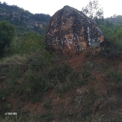 A large, weathered boulder dominates the scene, marred by white graffiti. The surrounding landscape features a hillside covered in sparse vegetation and a backdrop of a forested mountain range. The image captures a somewhat desolate, rural environment with signs of human impact on a natural feature. The bottom left corner displays a "Tecno" watermark.