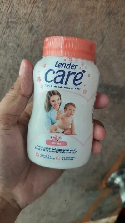 A close-up, eye-level shot shows a bottle of "tender care hypoallergenic baby powder" being held by a person's hand against a rustic wooden background. The bottle has a pink ribbed cap and a white body featuring a graphic of a woman and a baby, along with text detailing the product's benefits. The brand name "tender care" is prominently displayed in blue lettering. A pink oval label with "sakura" written on it is visible below the baby graphic, followed by text stating "Protects by helping keep your baby's skin comfortable and dry." Additional information such as "Gentle for sensitive skin," "0% Parabens & Silicones" is also present. The hand holding the bottle appears to be an adult's with fair skin and is positioned to obscure the lower-left quadrant of the bottle. The wooden surface behind the bottle displays a natural grain pattern. The location of this image is not specified and cannot be determined.
