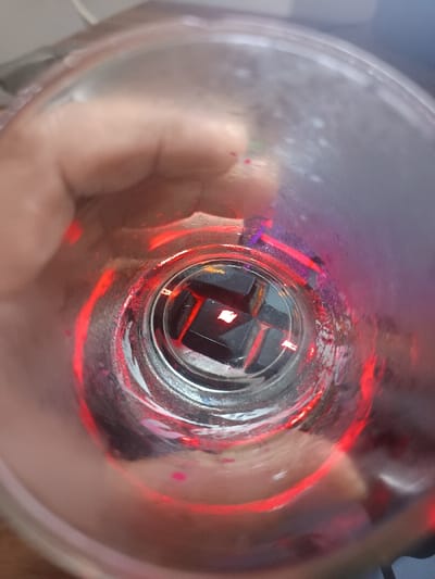 The image displays a top-down, interior view of a translucent, cylindrical container, likely a cup, held by a human hand. The container's inner surface exhibits dried residues of a red substance, with minor traces of purple or pink, and appears smudged. At the bottom of the container, several small, dark, cuboid objects are visible, consistent in appearance with mechanical keyboard switches. One of these switches is distinctly illuminated from within, emitting a bright reddish-orange light that strongly reflects off the interior walls of the container in radial streaks. A human hand, with light skin pigmentation, is visible on the exterior of the container, grasping it from the left side of the frame. The background external to the container is out of focus and non-descript, preventing specific environmental or geographic identification. The location in Ouderkerk aan de Amstel, Netherlands, cannot be determined from the visual evidence.