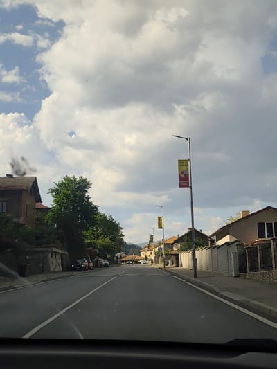 The image shows a street scene in Mesta, Bulgaria, taken from inside a vehicle looking down the road. The sky is mostly overcast with large cumulus clouds. The road is asphalt with a central white dividing line. On the left side of the road, there are parked cars, some buildings, and trees. On the right, there is a long concrete wall. Street lamps line the road on both sides, some with banners advertising the local soccer club in Bulgarian, "Бъди на стадиона" and "Подкрепи отбора". Further down the road, there are more buildings and trees. A group of people are near one of the parked cars on the left, appearing to interact. The vehicle's dashboard is visible at the bottom of the frame.