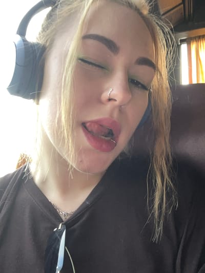 A close-up, tilted, and angled view captures a person with blonde hair, closed eyes, and red lipstick. The individual has a nose ring and is wearing headphones. They also wear a dark-colored shirt and a silver necklace. The background appears to be indoor, possibly near a window with blinds. The location, Prijevorac, Montenegro, is not visually apparent in the image.