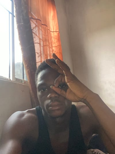 A close-up, eye-level shot captures a person of dark skin tone, likely male, looking directly at the camera. The image is oriented vertically, with the person's head facing towards what would be the right side of a standard portrait orientation. The individual wears a dark, possibly black, tank top or sleeveless shirt. Their left hand (viewer's perspective) is raised to their face, with two fingers extended in a V-sign gesture or partially obscuring their nose and forehead. Their eyes are dark and look directly into the lens.
The background consists of a brightly lit window on the left, partially covered by an orange or sheer brown curtain. The strong light from the window suggests daytime. Below the curtain, a blurry, bright outdoor scene is visible, with some faint dark lines suggesting window frames or bars. Above the person's head, a light-colored ceiling or wall is visible. The lighting creates highlights on the person's skin, particularly on their forehead, nose, and shoulders. No text is visible in the image.