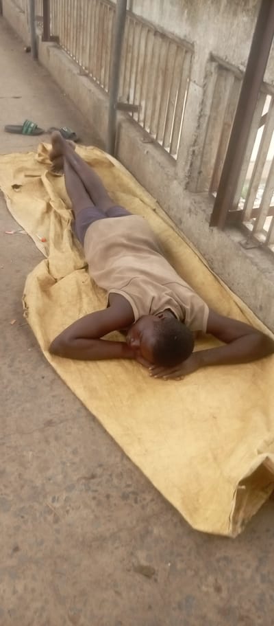 A young person lies on a yellow tarp-like material on a concrete surface. The person is wearing a tan shirt and darker shorts. The individual is lying on their back with their head resting on their folded arms. Their legs are outstretched. In the background, there is a metal railing and a concrete wall. There are sandals near the person's feet. The scene appears to be taking place outside. The location is indicated as Idemili, Nigeria.