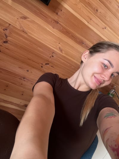 A medium close-up, high-angle selfie photograph depicts a young adult female smiling and looking directly into the camera. She has light skin, light brown hair pulled back, green eyes, and pink lips. She is wearing a short-sleeved, dark brown top. Multiple colorful tattoos are visible on her right forearm, including floral motifs. Her left arm is extended towards the camera, and a portion of a blue object, possibly a chair or fabric, is visible beneath her right arm. The background consists primarily of light brown wooden wall paneling with prominent grain and knots. In the upper left corner, a partial black rectangular object is visible. To the right, behind the subject's right shoulder, a white bottle with a green and white label (partially legible as "Fru...") is visible next to a silver faucet or tap. The scene is located in Доња Брезна, Montenegro.