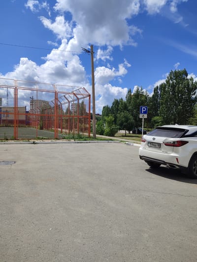 The image shows a street scene in the city of Lisakovsk, Kazakhstan, on a bright, partly cloudy day. In the foreground, a white Lexus RX 300 SUV is parked in a paved lot. The vehicle's license plate is visible and reads "kz 15 IRAA 10". Adjacent to the parking lot, there is a fenced sports court with orange metal posts and netting, and a green surface. In the background, there are residential buildings and tall green trees. A blue and white parking sign for disabled individuals is also visible. The sky is a vibrant blue with scattered white clouds.