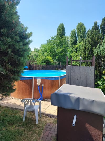 The image depicts an outdoor scene in Opatovice, Czechia, featuring a swimming pool and surrounding amenities. A round, above-ground swimming pool with bright blue water is visible, its wooden-patterned exterior supported by white poles. A white filter housing is attached to the pool wall, with blue hoses connected to it. To the right of the pool, a structure resembling an outdoor shower or changing enclosure is partially visible, constructed from dark grey, wood-textured panels. In the foreground, a white plastic chair sits on a paved area adjacent to a grassy lawn. A large, dark-colored cover is placed over a brown structure in the lower right quadrant of the image. The background is filled with lush green trees under a clear blue sky. No people are present in the scene.