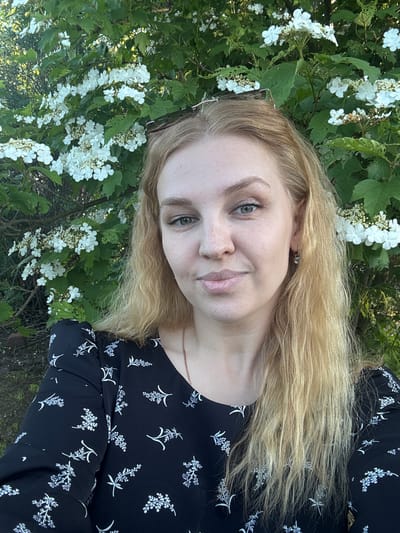 A frontal portrait shot shows a fair-skinned woman with long, blonde hair, grey-blue eyes, and light freckles, standing outdoors in Urmanets, Russia. She has sunglasses perched on top of her head. She is wearing a long-sleeved black garment adorned with a repeating white floral or leafy pattern. A thin gold-colored chain necklace is visible, along with a small, clear stone earring in her right ear. The woman looks directly at the camera with a slight, closed-lip smile. Behind her is a dense background of green foliage interspersed with numerous clusters of small white flowers, characteristic of a flowering shrub. The lighting suggests natural daylight.