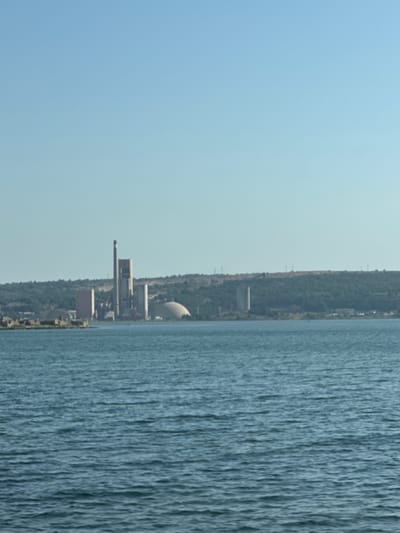 The image depicts a factory complex situated along a body of water, likely a river or sea, with a clear blue sky overhead. The orientation of the image is such that the factory appears to be on the left, and the water occupies the right side. The factory itself is an industrial facility with several buildings, including a tall, cylindrical structure that resembles a cooling tower, and a long, narrow building with a protruding element. There are also smaller, rectangular structures. The factory is surrounded by green vegetation, suggesting a rural or semi-rural setting. The water is rippled, indicating some movement on its surface, and its color is a deep blue-green. The scene suggests an industrial operation in proximity to a natural water source. The time of day appears to be daytime, given the bright sky, and the weather seems fair. There are no people visible in the image. The location is identified as Bages, France, which places this industrial site within that region. No text is discernible in the image.