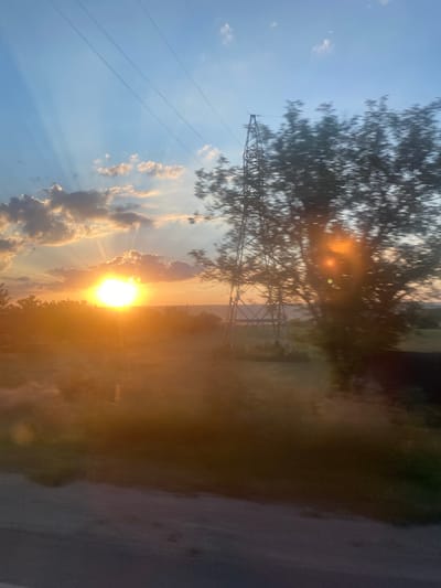 This image depicts a sunset scene near Balabanu, Moldova. The sun, a bright orb of light, is partially obscured by clouds, casting golden rays across the sky. In the foreground, a large transmission tower stands silhouetted against the vibrant sky, with power lines stretching across the frame. A blurry tree is visible to the right of the tower, its leaves blurred by motion. The landscape below is a mix of fields and trees, bathed in the warm, orange glow of the setting sun. A road or pathway is visible in the lower portion of the image, its surface blurred by motion. The overall impression is one of a serene, rural sunset with industrial elements.