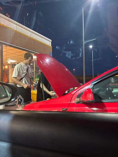In Sękocin Stary, Poland, two individuals are observed near a red car with its hood open, at night. The individual on the left, wearing a white t-shirt and black shorts with white trim, holds a clear plastic bottle of water. The second individual, dressed in black, appears to be attending to the car's engine. The setting suggests a public area, possibly a gas station, indicated by the building in the background featuring a yellow and brown facade with illuminated signage and an exterior light fixture. The overall scene is illuminated by artificial lighting, casting shadows and creating reflections.