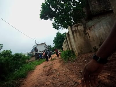 The image captures a scene on a dirt road in Amokpara, Nigeria. The road appears to be a path through a semi-rural or developing area, with lush green vegetation bordering one side and a dilapidated concrete wall on the other. In the background, modern buildings with white exteriors and dark accents are visible, suggesting a contrast between older and newer constructions. A group of individuals, appearing to be about five or six people, are congregated on the road. Some are standing, and one person is partially obscured by another. A pink motorcycle is parked near them. The sky is overcast and bright white. In the foreground, a hand with a black watch is positioned, with the wrist extending towards the camera. The overall lighting is subdued, indicative of an overcast day.
