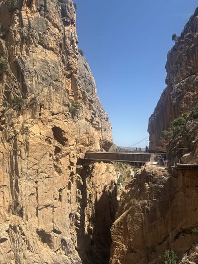 The image depicts a suspension bridge spanning a deep gorge with sheer rock faces on either side. The bridge, constructed with wooden planks and metal cables, appears to be a walkway or trail. Several people are visible on the bridge, walking and observing the surroundings. The scene is set in a natural environment characterized by rugged, rocky cliffs, with sparse vegetation growing from crevices. The time of day appears to be midday, as indicated by the bright sunlight casting strong shadows on the rock faces and the clear blue sky. The vastness of the gorge and the height of the bridge suggest a dramatic and potentially exhilarating experience for those traversing it. The terrain suggests a mountainous or canyon-like location, consistent with the context of Ardales, Spain, known for its dramatic landscapes and hiking trails like the Caminito del Rey. No visible text is present in the image.