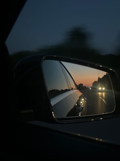 The image depicts a side-view mirror of a car, reflecting a road scene at dusk. The mirror shows a twilight sky with hues of orange and purple, indicating a sunset or sunrise. Trees line the road, and the headlights of an oncoming vehicle are visible, suggesting it is moving towards the camera. The reflection also captures the side of the car from which the photo is taken, including a portion of the car's bodywork. The overall impression is that of a journey taken during transitional light conditions. No specific location in Elsdorf, Germany can be identified from the visual information provided.