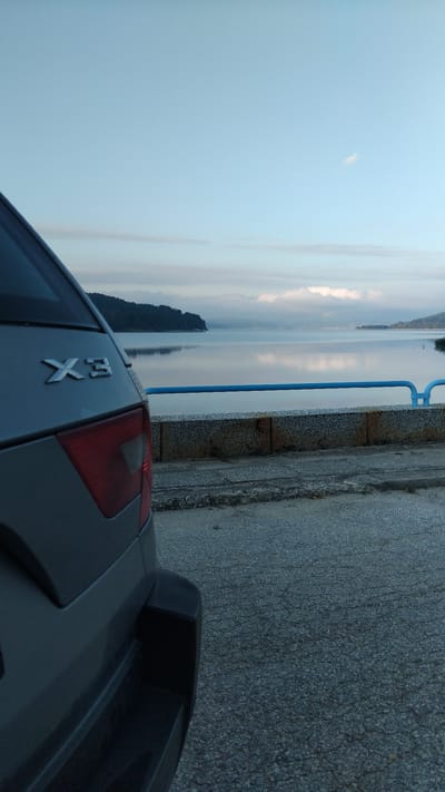 The image shows the rear left quarter of a silver BMW X3 parked on an asphalt surface next to a body of water. The vehicle is angled towards the water, with its taillight visible. In the background, there is a calm body of water reflecting the sky and a tree-lined shore. A blue railing runs along the edge of the water. The sky is overcast with some clouds, and the overall lighting suggests it is either early morning or late evening. The scene appears to be a scenic viewpoint, possibly in a coastal or lakeside area. The location is identified as Sv. Konstantin, Bulgaria, though there are no specific landmarks to confirm this within the image itself. There are no people visible in the image.