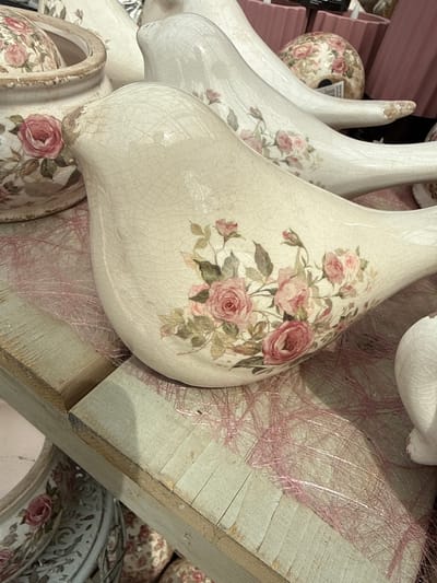 The image depicts a still life arrangement of ceramic decorative items, specifically birds and a pot, adorned with a floral pattern featuring pink roses. The main subject is a cream-colored ceramic bird with a crackled glaze and the rose pattern prominently displayed on its side. Behind it, a longer, white ceramic bird with a similar floral motif is partially visible, along with another round ceramic object decorated with roses. To the left, a ceramic pot, also decorated with roses, is seen. The items are placed on a wooden surface, possibly a shelf or table, with a delicate pink, possibly raffia or tinsel-like, material scattered around them. In the background, there are glimpses of pink cylindrical objects and other decorative items. The lighting suggests an indoor setting, possibly a shop or home. The image does not contain any people or indicate any specific location within the city of Fritzlar, Germany.