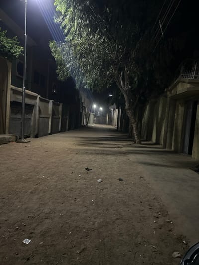 An unpaved dirt street, approximately 5 meters wide, extends into the distance under the night sky in Yalwa, Nigeria. The street is illuminated by at least three streetlights: a prominent, bright white LED lamp on a tall pole is positioned on the left in the foreground, and two additional lights are visible further down the street, casting long shadows.
On the left, a low concrete curb borders a textured concrete wall with multiple recessed panels, some appearing to be metal gates or doors. Behind this wall, a dark building with visible windows or openings rises. On the right, a multi-trunked tree with dense, dark green foliage stands prominently, casting shadows across the street. Beyond the tree, a beige-colored wall with vertical pilasters extends down the street, leading to obscured structures. Overhead, utility lines are visible spanning the street.
The dirt ground shows tire tracks and scattered debris, including small rocks and several dark, amorphous objects. A distinct white square object with a dark pattern is present on the ground in the lower-left foreground. No individuals, animals, or active movements are observed. The scene is static.
