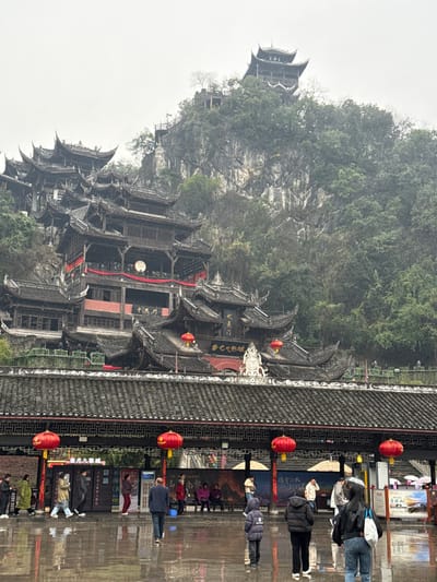 This image depicts a traditional Chinese architectural complex, identified as Chiyou Jiuli City (蚩尤九黎城), situated on a mountainous landscape in Pengshui Miao and Tujia Autonomous County, China. The scene is under an overcast sky, with the foreground ground visibly wet and reflective, indicating recent or ongoing rain.
In the background, a steep, densely vegetated mountain face rises, covered with trees and rocky outcrops. Multiple tiers of traditional Chinese buildings, characterized by dark, curved, multi-eaved roofs and dark wooden facades, are constructed directly into the mountainside. Red banners are draped across sections of these buildings, and a prominent large, circular white object is visible on one of the central structures. At the mountain's peak, a distinct, taller pagoda-like building with multiple stacked roofs is visible, accessible via a narrow path with railings.
In the mid-ground, at the base of the mountain, a substantial gatehouse or entrance structure with a broad, dark-tiled roof, decorative elements, and multiple red lanterns hanging beneath its eaves is positioned. Chinese characters, including "蚩尤九黎城," are prominently displayed on signage within this structure.
The foreground consists of a wide, wet, paved plaza. Approximately twenty-five individuals, including adults and children, are present. They are dressed in casual clothing, with many wearing jackets suitable for cool or wet weather. Several red lanterns are suspended from the eaves of the mid-ground structure, extending into the frame. People are observed walking across the plaza, standing in small groups, or sitting on benches positioned along the base of the entrance structure. Some individuals are holding umbrellas. No distinct interactions or specific actions beyond movement and resting are discernible.