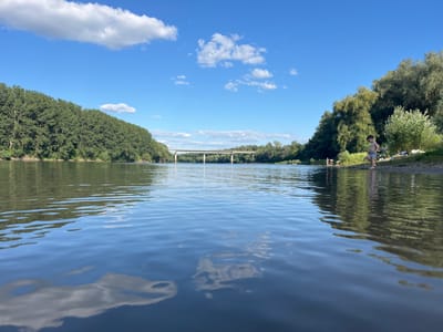 This is a low-angle, eye-level, full shot of the Nistru River in Vadul lui Vodă, Moldova, on a sunny day. The water occupies the foreground, reflecting the blue sky and white clouds, and ripples gently. Along the left bank, dense green trees line the river, extending into the distance. In the center of the image, a concrete bridge spans the river, with a few people visible on the riverbank on the right. The right bank is characterized by green vegetation and trees, with several individuals, including a young boy standing on the shore, interacting near the water's edge. The overall scene depicts a peaceful, natural setting with recreational activity.