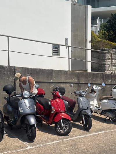 A line of scooters is parked along a concrete barrier. The scooters are white, gray, and red. A man wearing a white tank top and tan baseball cap bends over one of the scooters. A white building is to the left, and green foliage is at the top of the frame.