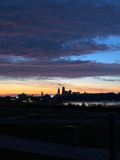 An evening photograph captures a twilight scene in Edgewater, New Jersey, United States of America. The sky is a gradient of deep blues, purples, and oranges, with clouds streaked with pink and blue hues. A silhouetted cityscape runs along the horizon, with several buildings and lights visible. A body of water, likely the Hudson River, lies between the foreground and the cityscape. The foreground consists of a park area with dark grass, paved pathways, and what appear to be benches or low walls. The lighting suggests a time shortly after sunset, with artificial lights beginning to illuminate parts of the park and the distant city. The overall atmosphere is serene and tranquil, with the natural light of the fading day meeting the emerging artificial lights of the urban environment.