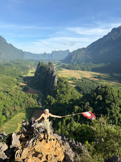A woman with blonde hair is standing on a rocky outcrop, holding a flag on a pole. She is wearing a black bikini top and olive green pants. She is barefoot. To her left, a dirt bike is parked on the rocks. Behind her is a vast, green landscape of mountains, valleys, and rice paddies, with a winding road visible in the distance. The sky is blue with some wispy clouds. The scene is set in the region of Naka, Laos.