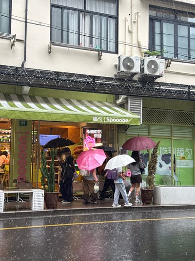The image captures a street scene in Xuhui District, China, during a downpour. Several people are using umbrellas to shield themselves from the rain as they stand on a wet sidewalk. A storefront with a green awning and large windows displays various products and signage. One sign, written in Chinese characters, reads "吃到舔勺" (chi dao tian shao), which translates to "eat till you lick your spoon," and also "Housemade YOGURT." Another sign for "希腊酸奶果昔" (Xila suannai guo xi) indicates "Greek yogurt smoothies."
Visible objects include air conditioning units mounted on the building's exterior, potted plants, wicker baskets, and white plastic storage containers. The street is slick with rain, and raindrops are visible throughout the frame, suggesting active precipitation. The scene appears to be during daylight hours, given the visibility, but the overcast weather due to the rain creates a somewhat muted atmosphere. The people's posture and use of umbrellas indicate they are likely waiting for something or passing by, with the weather dictating their current activity.