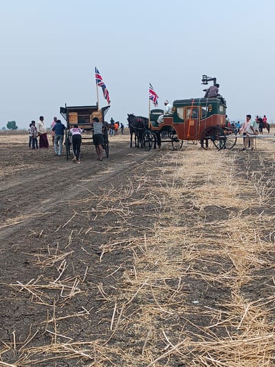 The image portrays an outdoor scene in a dry, barren field within Kottalapalli, India, under an overcast sky. Two prominent horse-drawn vehicles are central. On the left, a dark, open-topped wagon containing wooden crates is being propelled or adjusted by at least two men from its rear. A Union Jack flag is mounted on its front. To the right, an ornate, enclosed vintage-style carriage, predominantly brown and dark green with gold accents, is stationary. Two dark horses are harnessed to this carriage. A camera rig is mounted on the carriage roof, with one man seated next to it. Another man occupies the driver's seat. A second Union Jack flag is affixed to the front of this carriage. Approximately 10-15 individuals are dispersed throughout the scene. Several men are actively pushing or stabilizing the left wagon. Two men stand near the right side of the ornate carriage. In the distant background, additional individuals are visible, including one on horseback in a red and white uniform and another wearing an orange vest near a horse. The ground consists of dark, cracked earth sparsely covered with dry straw stubble, featuring a visible dirt track. Faint wind turbines and trees are discernible on the distant horizon. The overall arrangement suggests a film or theatrical production set.