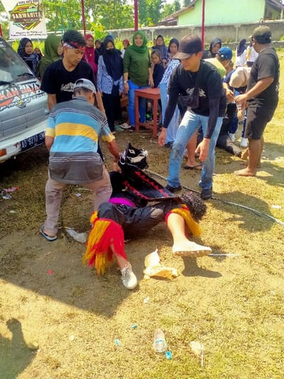 An outdoor scene in Celep, Indonesia, depicts a cultural performance on dry, patchy ground under bright sunlight. A performer, dressed in dark clothing with red and yellow fabric fringe around the lower legs and white sneakers, lies on their back/side. A thin metal rod pierces the performer's lower right leg, displaying minor bleeding. A stylized black and white hobby horse prop rests near the performer's upper body.
Three men are positioned around the performer: one in a blue, yellow, and grey striped polo shirt and grey pants is bent over, interacting near the performer's head; another in a black t-shirt, blue jeans, and black baseball cap stands leaning forward, observing; and a third in a black t-shirt and black shorts stands barefoot to the right, also observing.
Approximately 15 to 20 spectators, predominantly women in hijabs and children, observe from the background. They are positioned behind a white utility van, displaying "SHUKAK JAYA ABADI" branding and Indonesian license plate "AD 9210 SM," and behind a light-colored wooden table with red legs. A white banner with "SARWAHITA" text is visible on the left side of the frame, above the van. Residential buildings with red roofs and green trees line the distant background. Scattered plastic waste and debris are present on the ground.