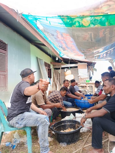 In Sangso, Indonesia, a group of men are gathered outdoors, seated on the ground and chairs under a makeshift tarp canopy. The canopy, made of assorted fabrics and banners, provides shade from the sun. Several men are actively engaged in food preparation, with some reaching into large plastic basins filled with what appears to be chopped food ingredients. One man in a black t-shirt and cap is gesturing towards the group, while others are focused on their tasks. There are several large black and blue basins, along with bags of what might be rice or flour. The environment is rustic, with a light blue-colored building in the background and scattered debris on the ground, suggesting a casual outdoor gathering or event.