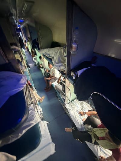 The image depicts the interior of a crowded train carriage, likely a sleeper class compartment, with people occupying multiple berths. The scene is dimly lit, with overhead fluorescent lights providing some illumination. Several individuals are visible, some sitting on their berths and others lying down. Bedding, consisting of white sheets and blankets, is spread across the berths. A water bottle is attached to the wall of the carriage. The overall environment suggests travel during nighttime. The presence of multiple passengers in close proximity and the arrangement of berths are characteristic of Indian Railways' sleeper class compartments. While the image itself does not provide definitive proof of the location being Mathura, India, this type of train carriage is commonly found on routes serving this city.