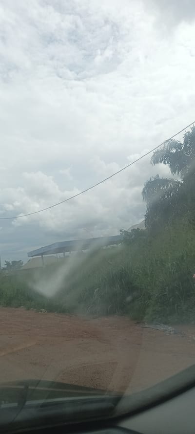 The image shows a view from inside a vehicle, looking out at a scene near Umuoye, Nigeria. A dirt road is visible in the foreground. Beyond the road, there is tall grass and vegetation, with a structure that appears to be a building or shelter. The sky is overcast with heavy clouds. A power line stretches across the upper portion of the frame. The foreground is partially obscured by a smudged or wet area on the windshield.