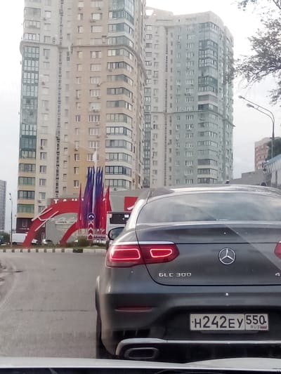 The image shows a street view in Gol'yovo, Russia, with a Mercedes-Benz GLC 300 car in the foreground. The car is facing away from the camera and is on a road. In the background, there are two tall residential buildings with many windows, and a large red arch structure decorated with flags. There are also streetlights and trees visible. The sky is overcast.