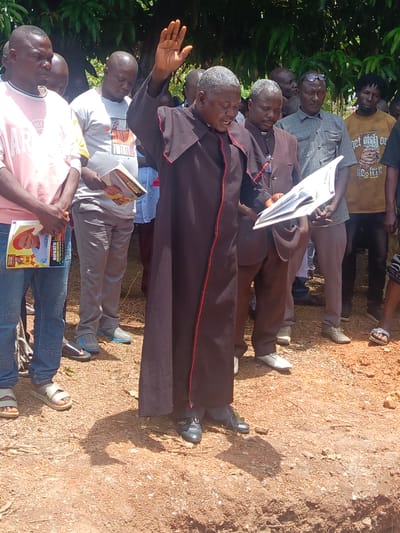 A man in a black robe with red trim stands on a dirt ground with his right hand raised, appearing to bless the surrounding group of people. He holds a white booklet in his left hand. Another man in a brown suit and clerical collar stands next to him, also holding a booklet. The people are dressed in casual clothing. Some individuals in the crowd hold pamphlets with a picture of a young boy and the name "RIFKATU KUCHA" printed on them. The environment suggests an outdoor setting, possibly a burial site, in Ninzo South, Nigeria.