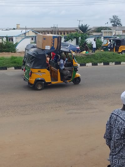 This media file captures a scene in Oja Oba, Oko Erin, Nigeria. The primary focus is a yellow and green auto-rickshaw, or "Keke," overloaded with goods. On top of the Keke are several large cardboard boxes, one prominently displaying a blue and white logo with text that includes "DELUXE". Inside the Keke, a man is visible driving, and another person is seated. The Keke is on a paved road, with a strip of grass and a sidewalk with black and white border stones alongside.
In the background, buildings with light-colored walls and dark roofs are visible, some with windows and doors. There are also trees and vegetation, suggesting an outdoor, possibly semi-urban or commercial area. A tall pole with wires runs across the top of the frame. To the right, another Keke is parked or moving, and a dark-colored car is partially visible.
Several people are present in the scene. Some appear to be standing or walking near the roadside. One man in a white shirt and blue trousers is standing in the middle ground. Further to the right, the back of a person wearing a patterned garment and a white head covering is visible in the foreground, looking away from the camera. The sky is overcast, indicating a cloudy day, likely mid-morning or afternoon. The overall impression is one of everyday transportation and commerce in a Nigerian setting.