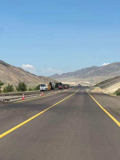 This image depicts a construction scene on a highway with mountains in the background. The highway is paved with asphalt and marked with yellow lines. Several trucks, likely involved in road construction, are present on the side of the road, along with traffic cones. The surrounding environment is arid, with dry, rocky hills and sparse vegetation. The sky is clear and blue. The scene suggests ongoing road maintenance or expansion work in a remote, mountainous region, possibly in or near the city of Alqabas, Kazakhstan.