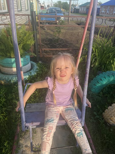 A young blonde girl with braided hair is sitting on a purple and red swing set in a yard. She is wearing a light purple t-shirt and light-colored pants with a cat pattern. Her face is contorted in a grimace, with her mouth open and teeth showing. The swing set frame is visible, with a wooden seat. In the background, there is a fence, overgrown green plants, and several repurposed tires painted in teal and cream colors, stacked and used as planters. Beyond the fence, a blue pickup truck is visible, parked near some houses. The scene appears to be in a rural or semi-rural setting, possibly in the village of Maltsevo, Russia, given the style of the houses and the overall environment. The lighting suggests it is daytime, likely late afternoon or early evening due to the shadows.