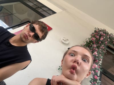 A low-angle, upward-facing shot captures two individuals posing for the camera, set against a light-colored ceiling or overhang. On the left, a female subject with light-colored hair makes a playful "duck face" with puckered lips and wide, open eyes. She wears a dark top and a ring on her right hand. To her right, a male subject is visible, wearing dark sunglasses that obscure his eyes and a dark, possibly black, collared shirt. He has short, dark hair and light stubble. A small silver hoop earring is visible on his left ear.

The background primarily features the light ceiling. Along the upper left, a lush garland of artificial pink roses and green leaves drapes, adding a decorative element. In the center, a small, circular, white light fixture is affixed to the ceiling. On the right side, behind the male subject, there's a dark window frame or architectural structure reflecting an outdoor scene with a cloudy sky and some circular patterns, possibly from another building or overhead structure. A small red rectangular object is also visible near the male's head on the ceiling.

The image appears to be a casual selfie or photograph taken from a very low vantage point, possibly from below a table or a railing. The overall lighting suggests it is daytime, with the male's sunglasses implying bright conditions outdoors. The artificial floral decor and architectural elements suggest a setting like an outdoor cafe, restaurant patio, or a covered walkway in an urban environment. No visible text is present in the image. This scene is located in Songjiang District, China.