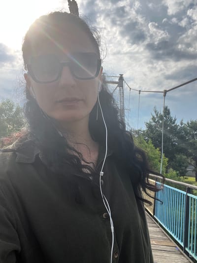 An outdoor selfie captures a person with long, dark, wavy hair, wearing a dark green, ribbed or corduroy long-sleeved shirt with prominent buttons. They are also wearing dark, square-framed sunglasses and white wired earbuds. The person is looking directly towards the camera, with a neutral or slightly smiling expression, though a prominent lens flare from the sun obscures part of their face.
The scene is set outdoors on what appears to be a bridge or elevated walkway, potentially in Kamianytsia, Ukraine. In the background, a cloudy sky with scattered white clouds is visible, illuminated by bright daylight. To the right, elements of a wooden bridge structure with distinctive blue metal railings are clearly seen, alongside lush green trees in the mid-distance. Overhead, suspension bridge cables and towers are discernible against the sky. The bright sun, positioned behind and slightly above the subject, indicates it is daytime, likely morning or late afternoon given the strong light angle. No visible text is present in the image.