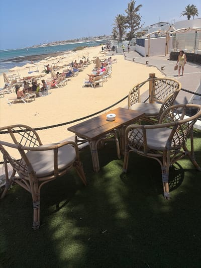 This bright, sunlit image captures a vibrant beach scene, likely at a resort in La Oliva, Spain. In the foreground, a patch of artificial green grass hosts several rattan-style outdoor dining sets, complete with chairs featuring white cushions and wooden tables. Strong shadows from overhead foliage indicate bright daylight. A small white, cylindrical object rests on one of the tables.

A thick, dark rope acts as a barrier, separating this grassy area from a wide, sandy beach that stretches into the mid-ground. Numerous beachgoers are visible, scattered across the sand, many relaxing on lounge chairs under cream-colored umbrellas. Further along the beach, white, modern-looking buildings with balconies line the shore, interspersed with tall palm trees. One person in a yellow top and shorts is seen walking on a wooden boardwalk near these buildings.

In the background, the clear, turquoise-blue ocean meets a vast, light blue sky. The water appears calm, and a distant coastline or landmass is faintly visible on the horizon. The overall scene conveys a sense of leisure and vacation under clear, sunny weather. There is no visible text.