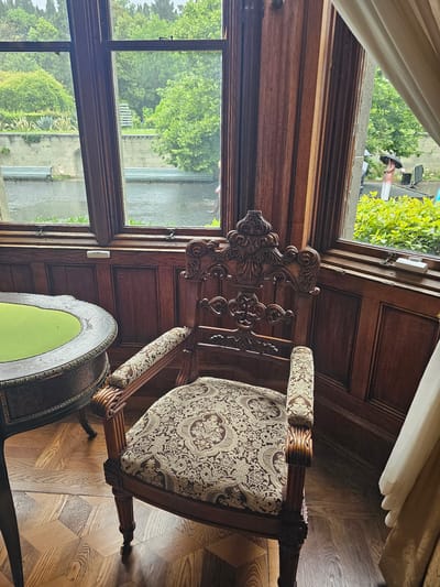 The image shows a room with an ornate wooden chair and table situated near a window. The room appears to be within a building located in Алупкинский городской совет, Ukraine, suggested by the view through the window. The window reveals an outdoor scene featuring trees, greenery, and what seems to be a waterway.
The chair is richly decorated with carved details and patterned upholstery. Next to it is a table with a green-colored top. The room is paneled in dark wood, adding to the traditional or historical atmosphere. The parquet floor features a geometric pattern.