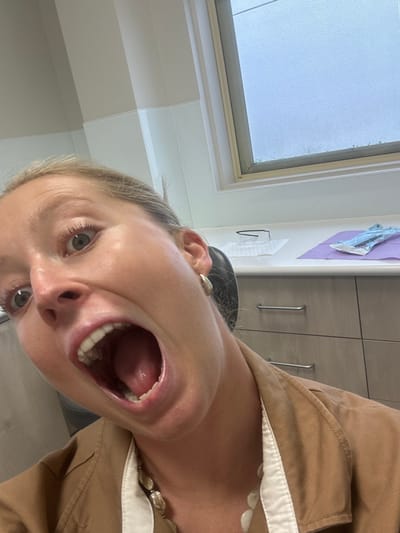 A woman is angled in the frame, facing downward. Her mouth is open wide. Behind her are bathroom cabinets with silver handles. A small, frosted window is visible above and to the left, showing an obscured view of the outside. The woman wears a tan shirt with white trim, and a pearl necklace. A hook hangs on the wall next to the cabinets.