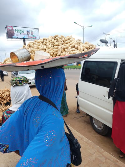 This media file captures a street scene in Oja Oba, Oko Erin, Nigeria. The central subject is a woman balancing a large tray of peanuts on her head. The tray is overflowing with the shelled nuts, and a large metal can and a white plastic container are visible among them. She is wearing a vibrant blue garment adorned with gold sequins.
To her left, another person, also wearing a head covering, is partially visible. Next to them, a pile of what appears to be dried root vegetables or snacks is stacked. In the background, a white van is parked, with its rear door open and a person in a red skirt standing beside it. Another person in dark clothing is seen walking away from the camera in the distance.
Above the scene, a large billboard with the text "RENEWED Hope" and images of three men is prominently displayed against a cloudy sky. The overall impression is that of a bustling market or roadside stall, likely during the daytime, with overcast weather. The focus on the woman carrying the peanuts and the surrounding goods suggests a commerce-related activity. The image is taken from a slightly low angle, emphasizing the weight of the peanuts on the woman's head.