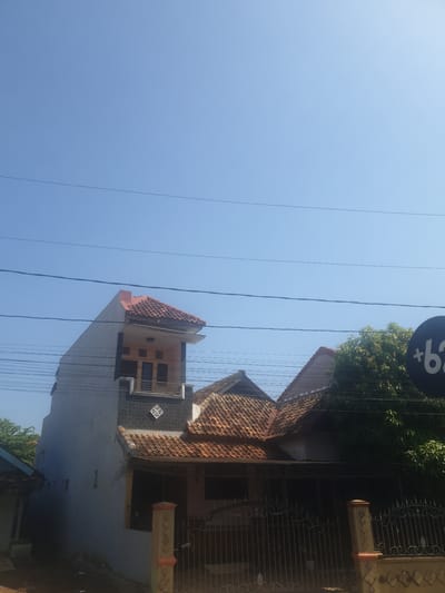 The image shows a residential area with two houses and a clear blue sky. One house is taller and appears to be a multi-story building with a tiled roof and gray exterior walls. It has several windows and a balcony. The other house is smaller, with a lower tiled roof and a visible fence in front. There are several electrical wires running across the sky, and a black circular sign with white text "+62" is visible on the right side of the image. Lush green trees are also present, partially obscuring one of the houses. The scene depicts a typical street in a residential neighborhood.